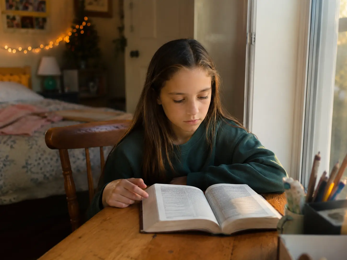A young girl reads an open Bible alone at a wooden table by a window in the warm afternoon light of her bedroom.