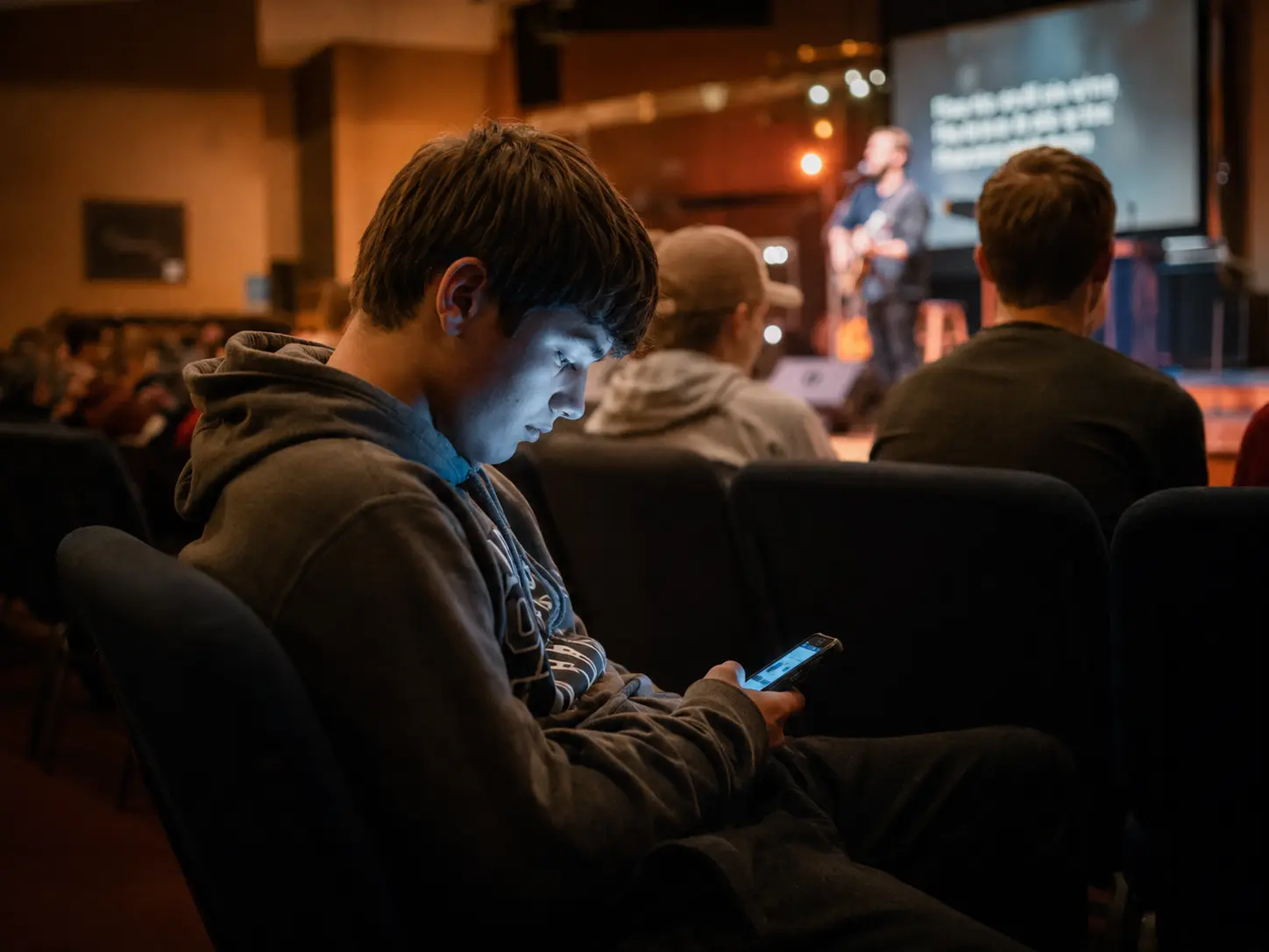 A teenager sits absorbed in his phone during a church service, his face lit by the cold blue glow of the screen while warm stage lights and a worship leader are visible behind him.